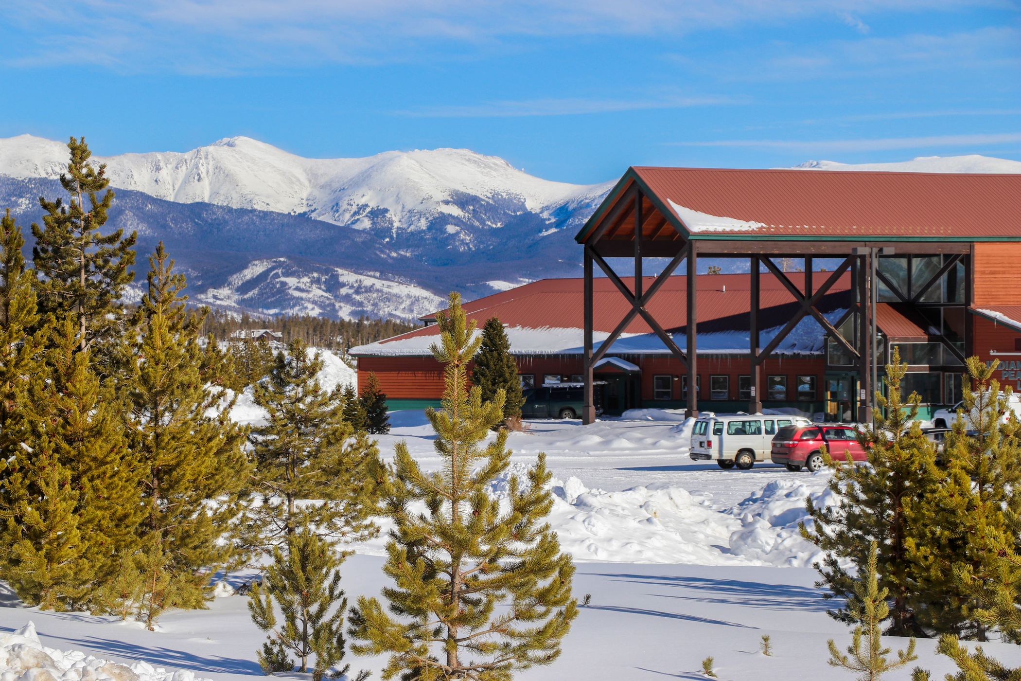 The front exterior of indian peaks lodge, parking lot covered in a thin layer of freshly plowed snow, with snow-capped mountains in the background.