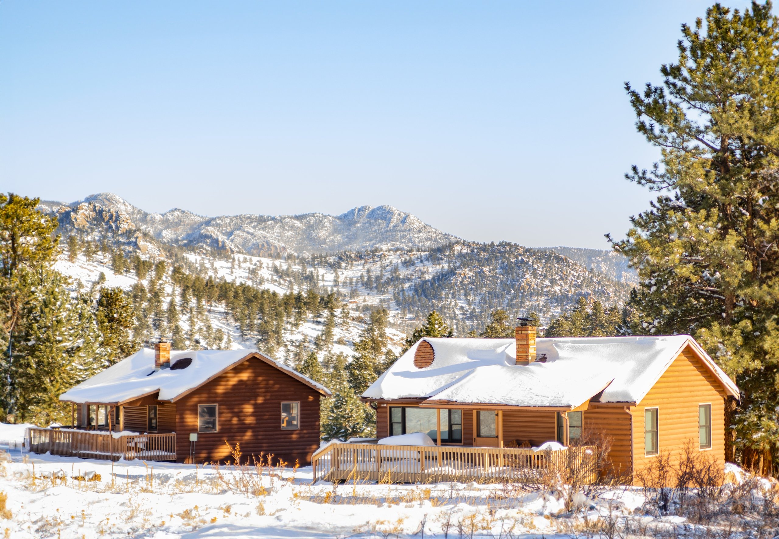 Cozy cabins, roofs covered in snow, looking out at a forested mountainside on a sunny winter day.