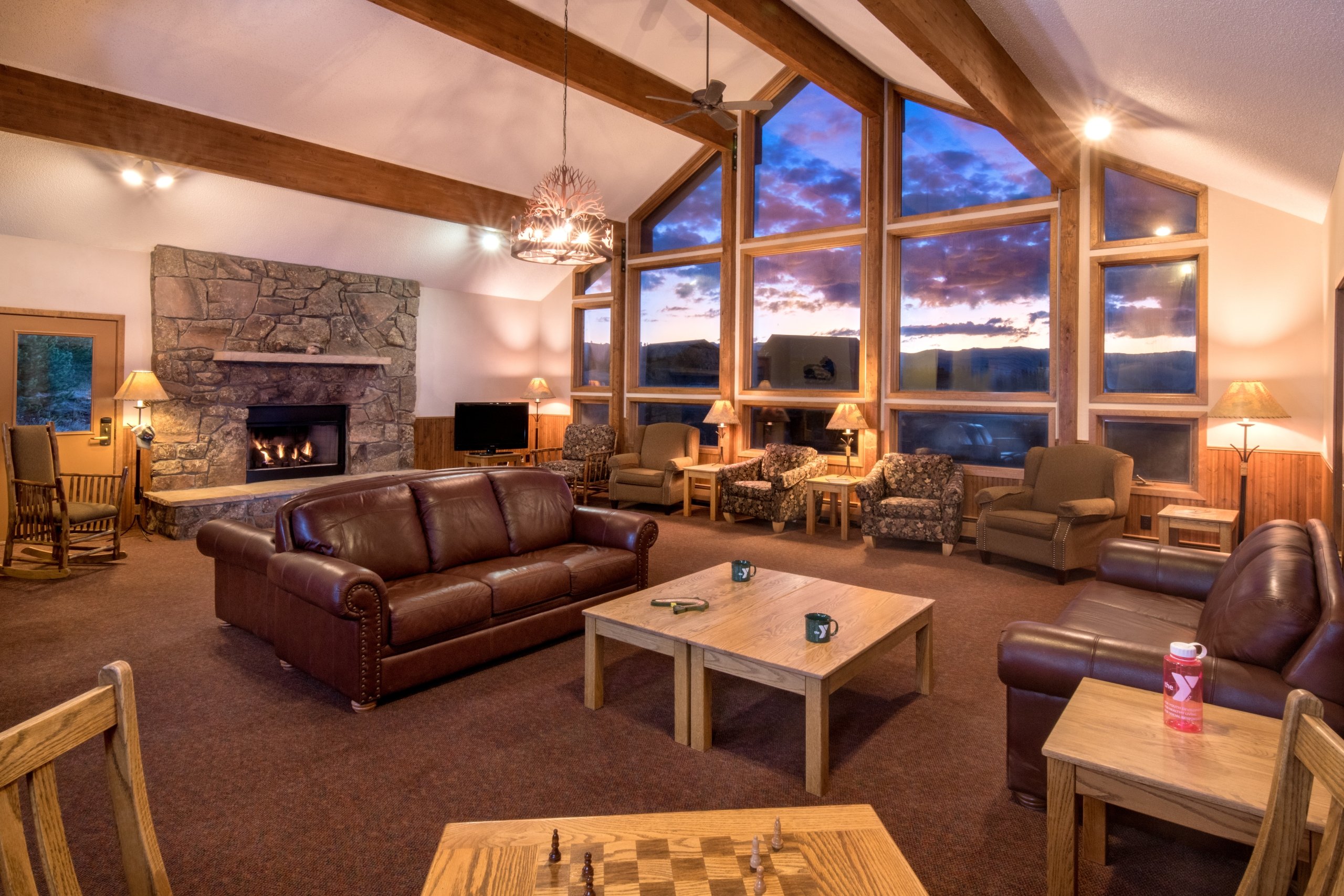 The living room of an 8 bedroom cabin, fully furnished with fireplace, 3 couches, 8 chairs, and a large window wall peering out at a sunset lighting up the continental divide.